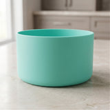 A matte mint green cylindrical bowl or planter sitting on a light beige marble countertop with a blurred kitchen background.