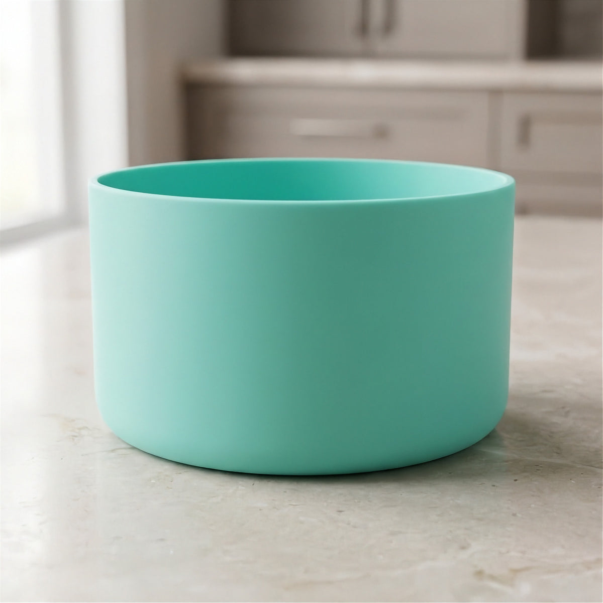 A matte mint green cylindrical bowl or planter sitting on a light beige marble countertop with a blurred kitchen background.