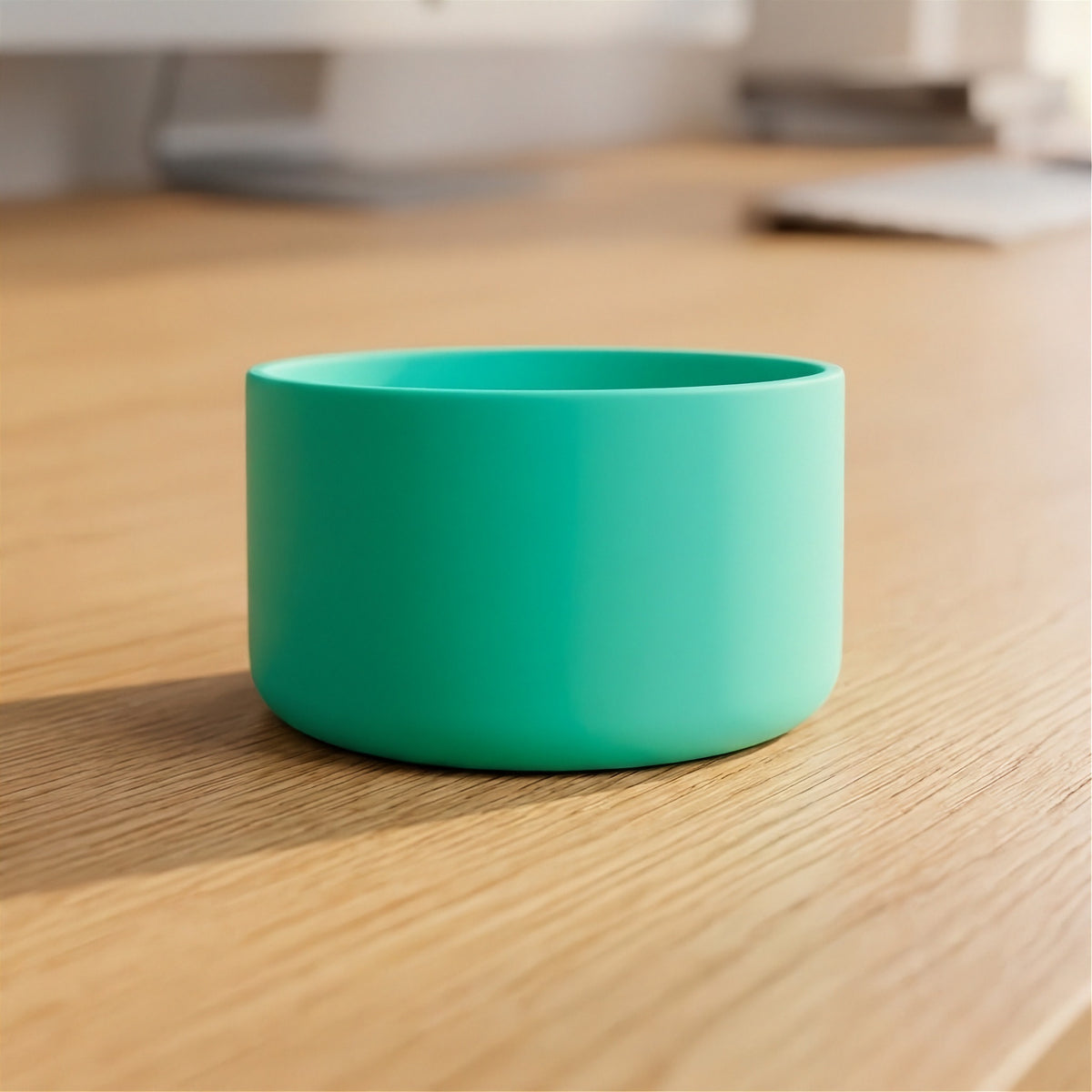 A bright teal-green cylindrical cup or small bowl with a matte finish, sitting on a light wood desk with a blurred office background.