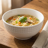 A white textured ceramic bowl filled with noodle soup, garnished with cilantro, carrots, and green onions, set on a wooden table next to a folded napkin.