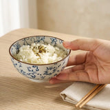 A hand holds a small white ceramic bowl decorated with blue floral patterns, filled with white rice topped with a sprinkle of seaweed, on a light wood table next to chopsticks and a napkin.
