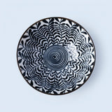 A round ceramic plate with a black and blue geometric pattern, featuring a central spiral design and radiating leaf-like shapes, set against a solid light blue background.