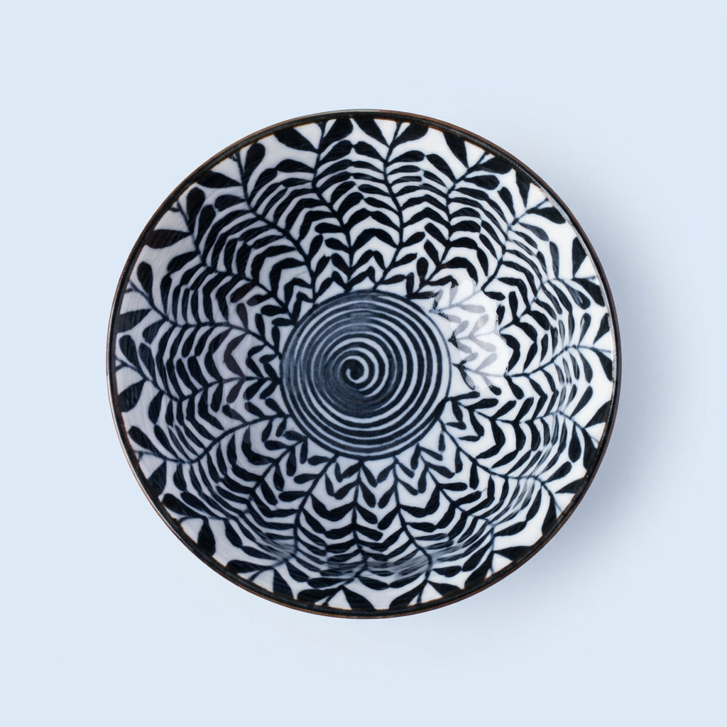 A round ceramic plate with a black and blue geometric pattern, featuring a central spiral design and radiating leaf-like shapes, set against a solid light blue background.