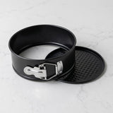 A black springform pan with a silver metal latch, shown next to its matching black textured lid on a light gray marble surface.