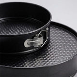 Close-up of a black springform pan with a textured non-stick surface and a silver metal clasp, resting on a matching round pan against a light gray background.