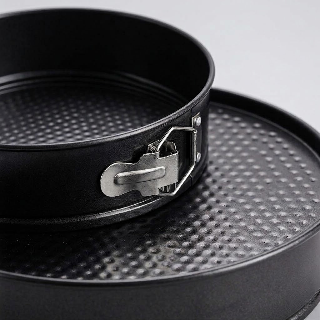 Close-up of a black springform pan with a textured non-stick surface and a silver metal clasp, resting on a matching round pan against a light gray background.
