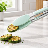 Light mint green silicone-tipped kitchen tongs with stainless steel arms lifting grilled zucchini slices on a light marble countertop in a bright kitchen.