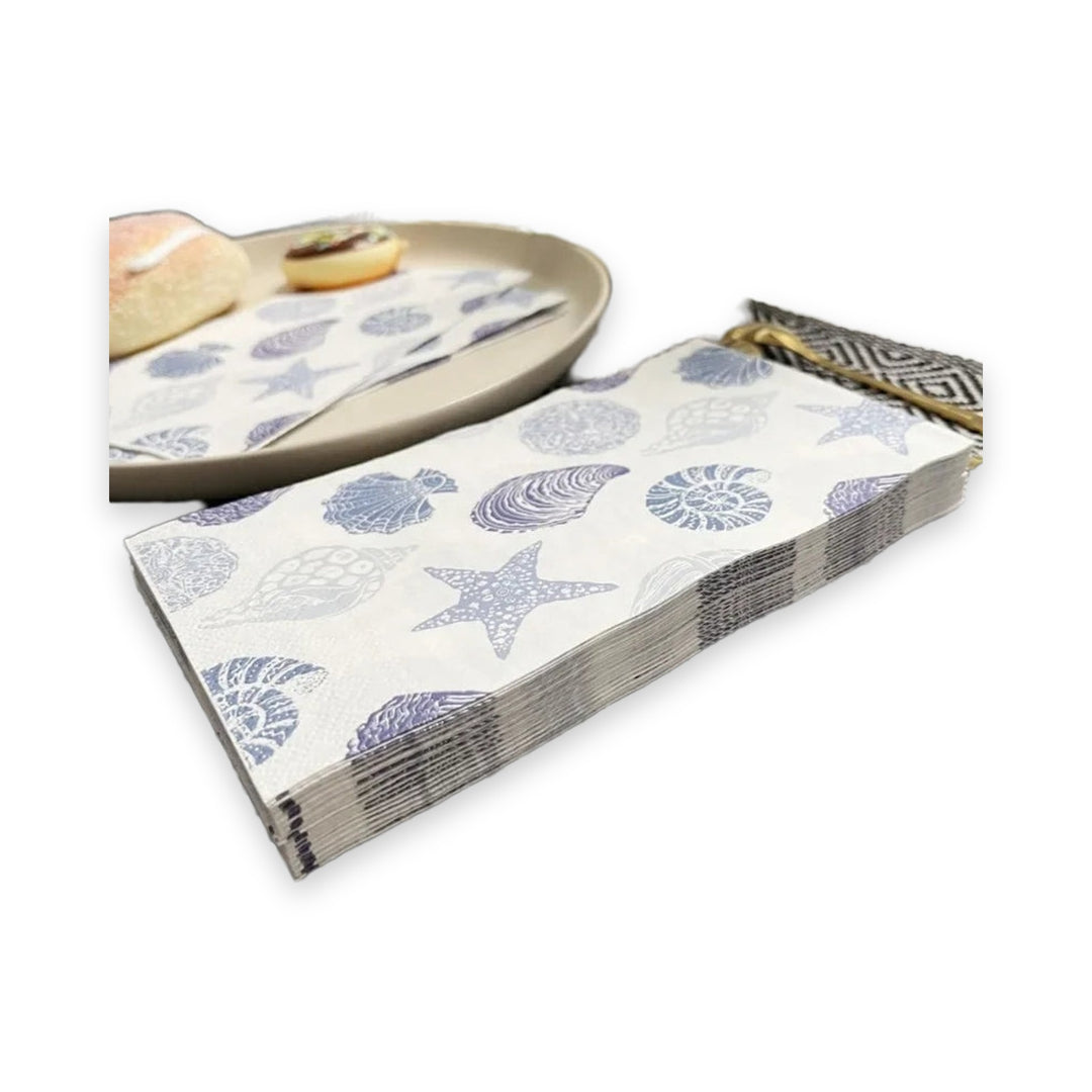 A stack of paper napkins with a blue and gray seashell and starfish pattern, shown alongside a matching napkin on a beige plate with bread, on a white background.
