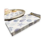A stack of paper napkins with a blue and gray seashell and starfish pattern, shown alongside a matching napkin on a beige plate with bread, on a white background.