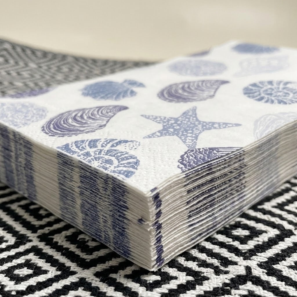 A stack of white paper napkins printed with blue seashell and starfish motifs, resting on a black and white patterned rug.