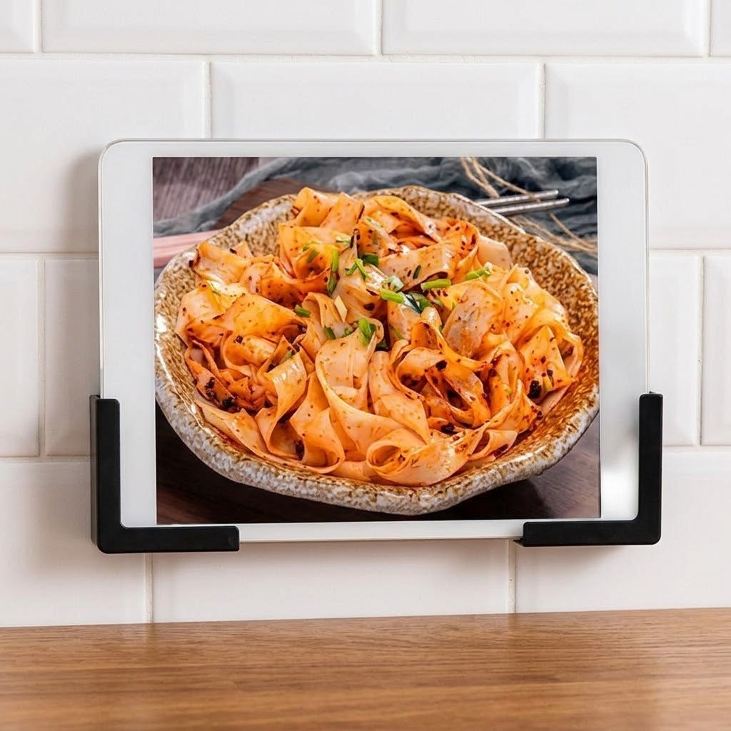 A black magnetic tablet holder mounted on a white subway tile backsplash above a wooden countertop, holding a tablet with a photo of spicy pasta on screen.