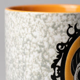 Close-up of a ceramic mug showing a speckled off-white exterior, a bright orange interior, and a black decorative scroll motif over a yellow section.