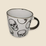 A white ceramic mug with a black rim and handle, decorated with a repeating pattern of black and white skulls, set against a solid beige background.