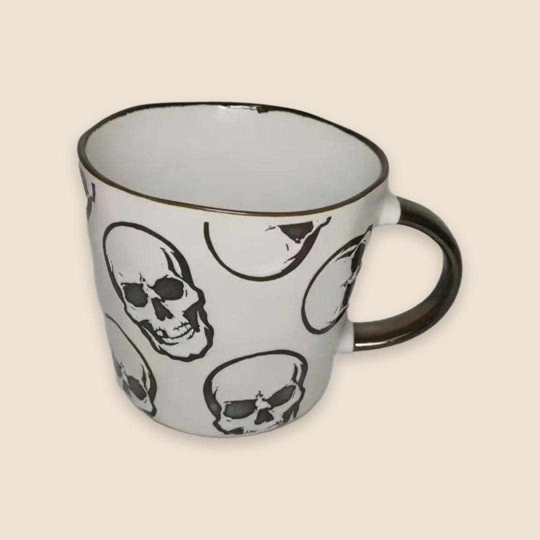 A white ceramic mug with a black rim and handle, decorated with a repeating pattern of black and white skulls, set against a solid beige background.