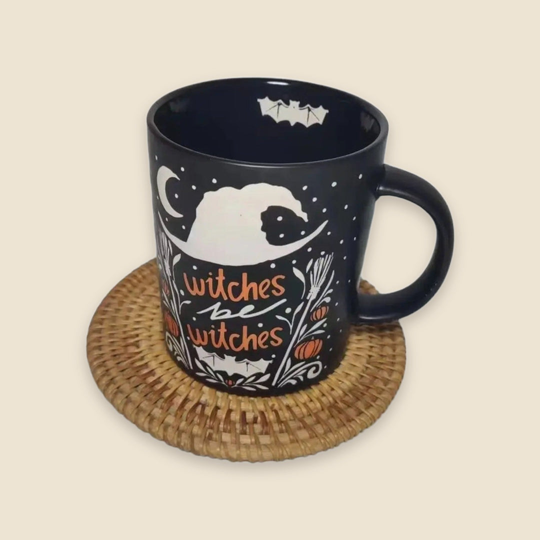 A black ceramic mug with a white witch-themed illustration and orange text reading 'witches' and 'witches', sitting on a round woven straw coaster against a plain beige background.