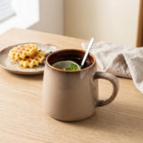 A ceramic mug with a warm brown and beige glaze sits on a wooden table, filled with a dark beverage and a lime slice with mint. A spoon rests inside the mug, with a plate of waffles and a beige cloth in the softly lit background.