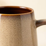 Close-up of a beige speckled ceramic mug showing its brown glazed rim, dark interior, and partial handle against a cream background.