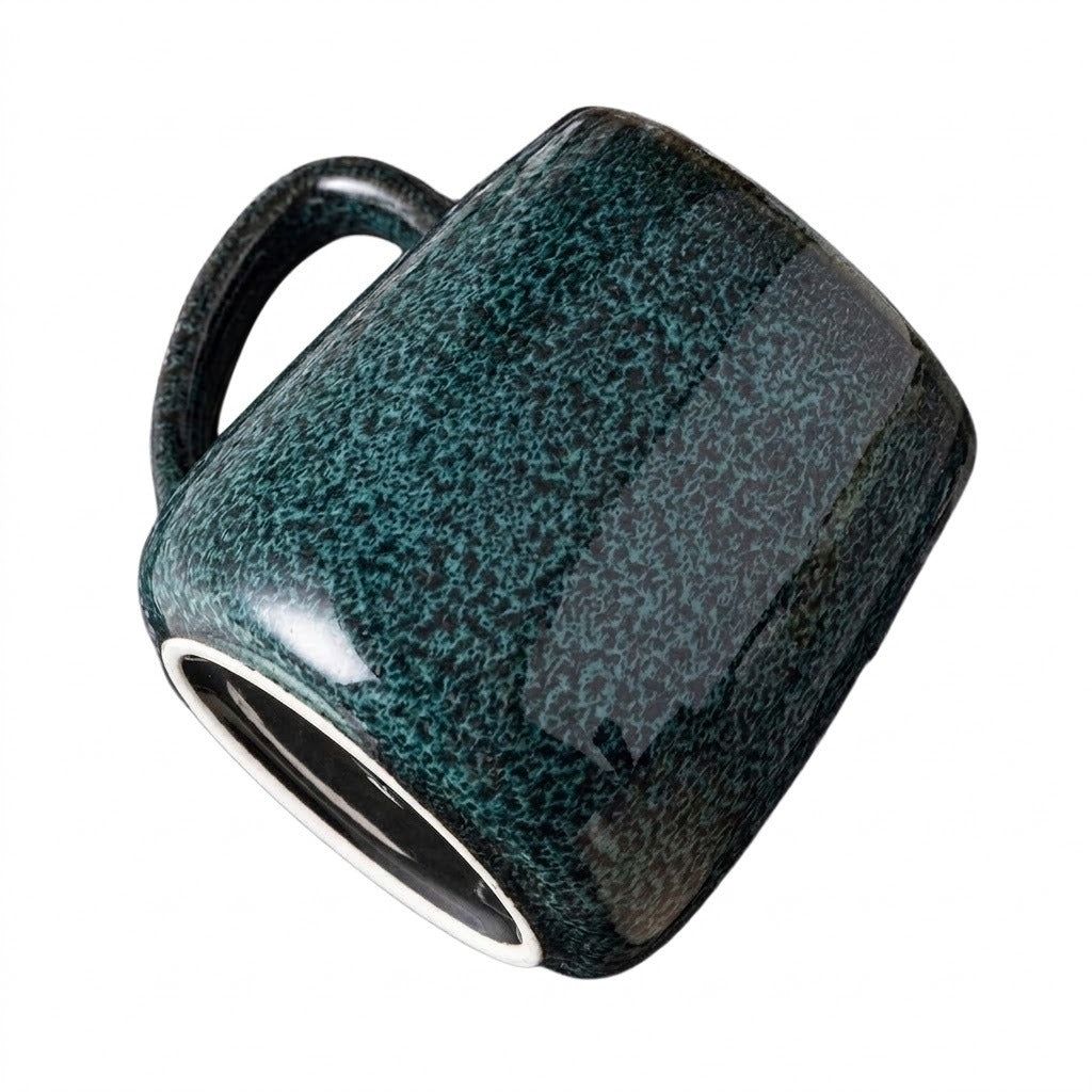 A dark teal speckled ceramic mug tilts diagonally against a white background, revealing its handle and white foot ring.