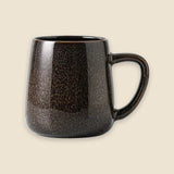 A dark brown ceramic mug with a speckled, textured glaze and a C-shaped handle, shown against a solid light beige background.