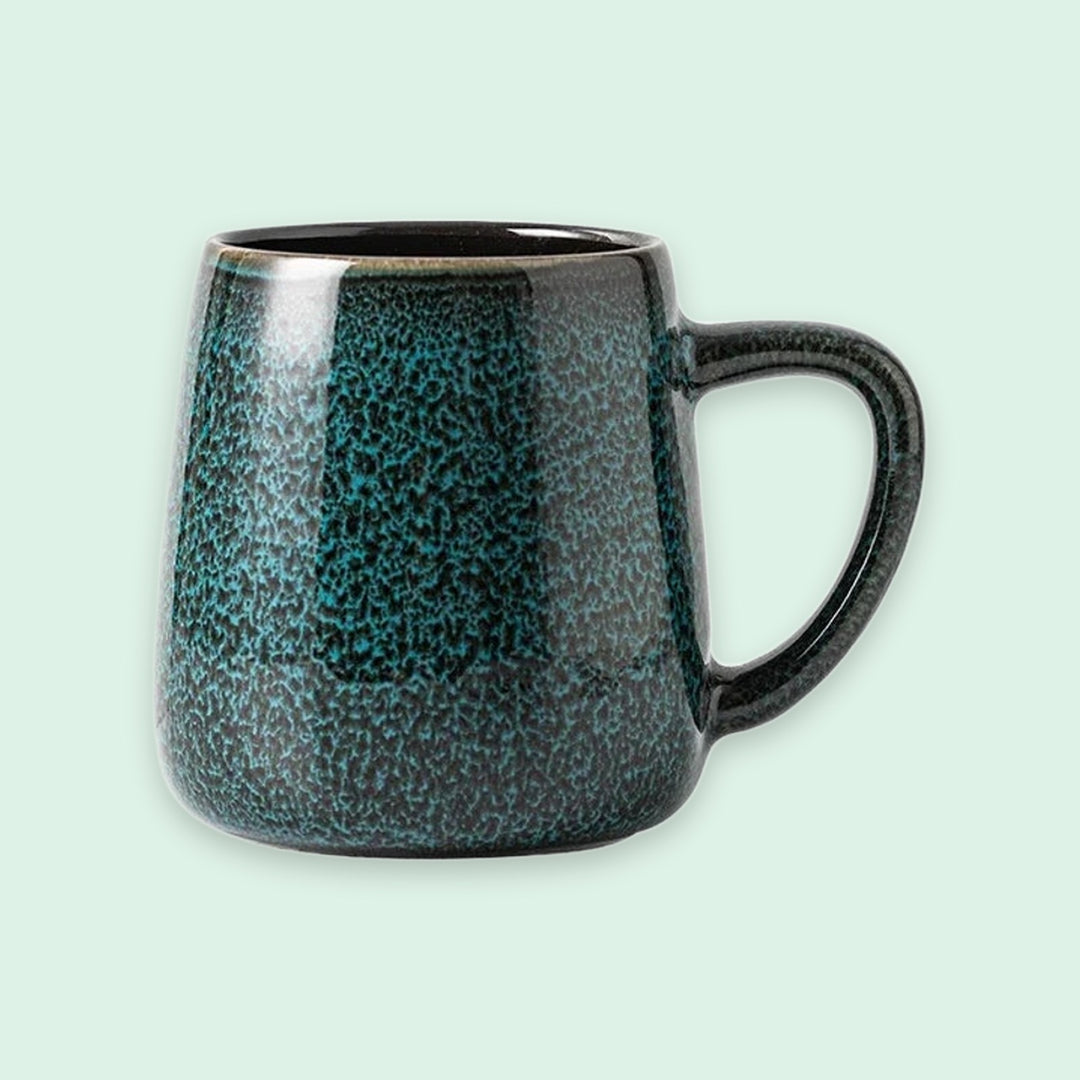 A ceramic mug with a textured, speckled dark teal and black glaze, shown against a solid pale mint green background.