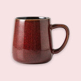 A glossy reddish-brown speckled ceramic mug with a dark interior and curved handle stands against a pale pink background.