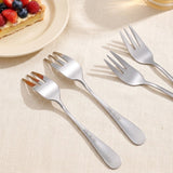 Four stainless steel dinner forks arranged diagonally on a beige textured tablecloth, with a partially visible slice of fruit tart on a plate in the upper left corner.