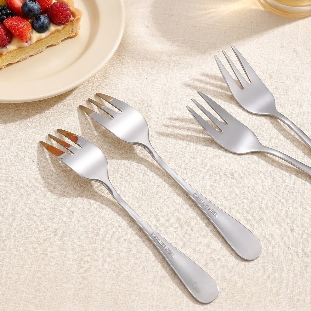 Four stainless steel dinner forks arranged diagonally on a beige textured tablecloth, with a partially visible slice of fruit tart on a plate in the upper left corner.