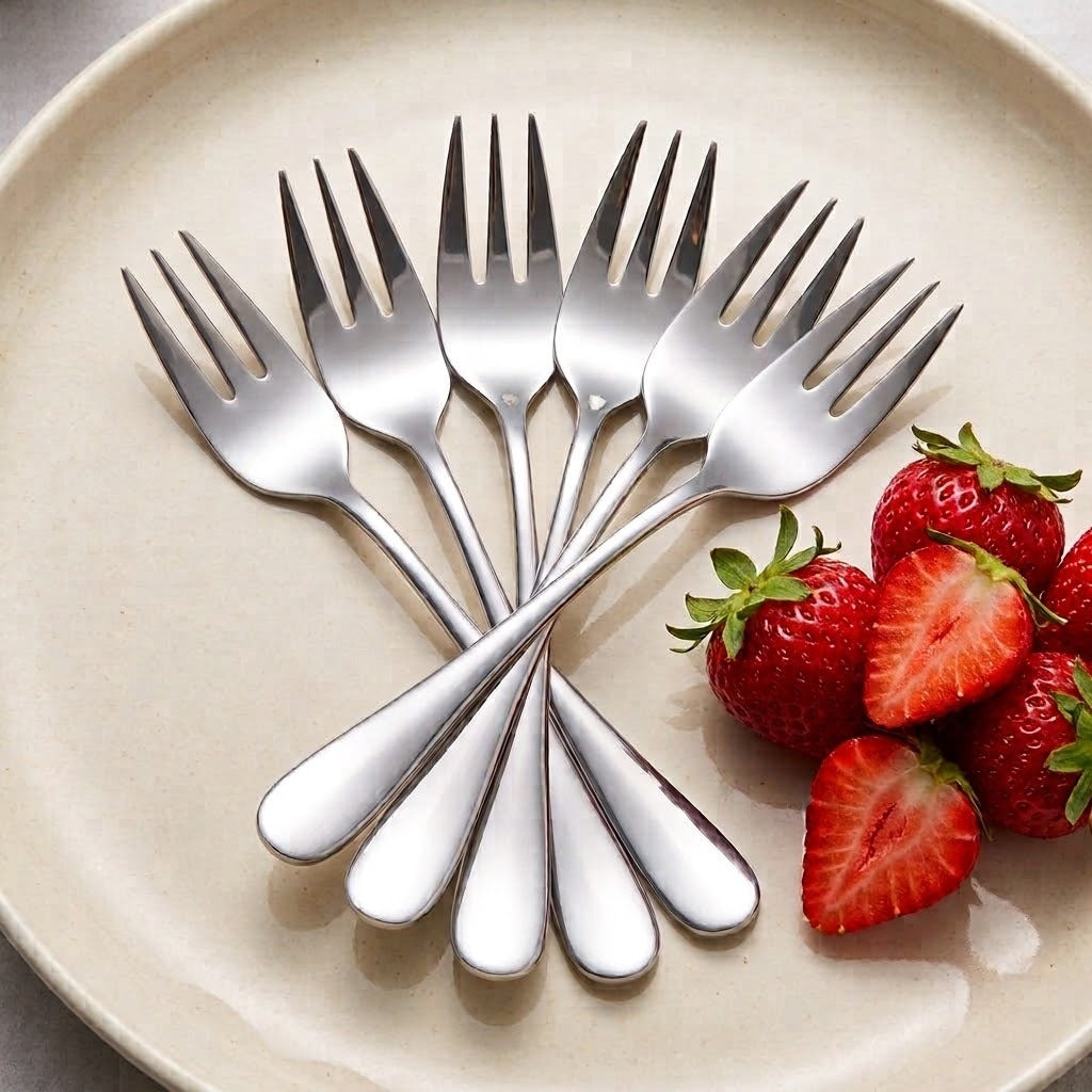 Six stainless steel dinner forks arranged in a fan shape on a beige ceramic plate, with fresh sliced strawberries placed on the right side.