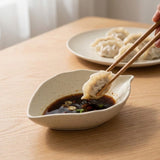 A close-up of a hand holding chopsticks with a dumpling above a small speckled ceramic dipping bowl filled with dark sauce on a wooden table, with a plate of dumplings blurred in the background.