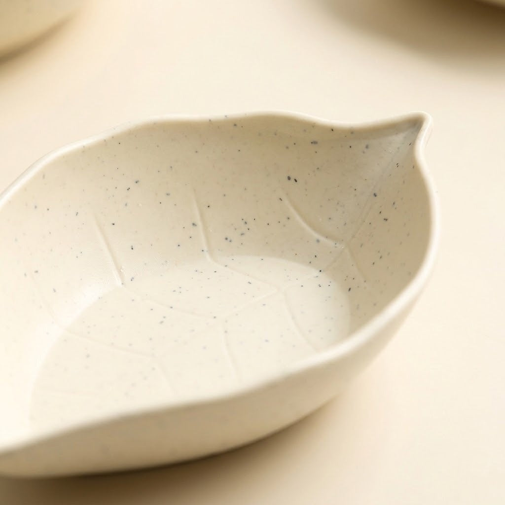 Close-up of a speckled cream leaf-shaped dish with raised vein details on a pale beige surface.