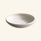 A shallow round ceramic bowl with a speckled off-white glaze and a slightly darker rim, shown on a plain light beige background.