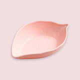A leaf-shaped ceramic dish in a speckled light pink and peach color, shown on a solid pale pink background.