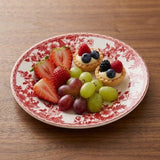 A white plate with red floral patterns holds strawberries, grapes, and two berry tartlets on a wooden table.