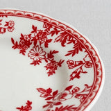 A close-up shows a white plate with red floral decoration and a patterned rim resting on light grey fabric.