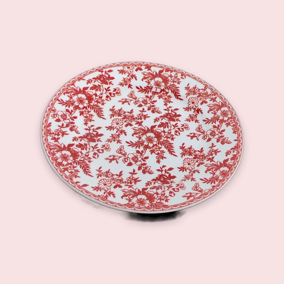 An oval ceramic plate featuring a red floral toile pattern on a white background, set against a solid pale pink backdrop.