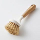 A dish brush with a wooden handle, white head and dense tan bristles lies diagonally on a white surface.