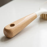 Close-up of a dish brush's wooden handle and hanging hole on a pale surface, with its white head blurred behind.