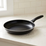 Empty black frying pan with a silver rim and black handle rests on a pale countertop against white tiles.