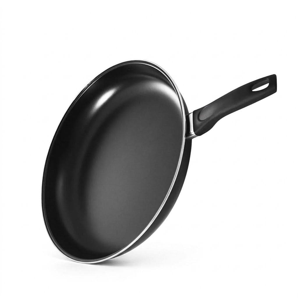 Black frying pan tilts upright against a white background, showing its smooth interior, silver rim, and handle with hanging hole.