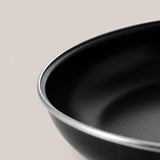 Close-up of a black frying pan shows its textured interior and silver rim against a pale beige background.