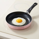 A pink frying pan with a black interior and handle holds a fried egg on a cream stovetop.
