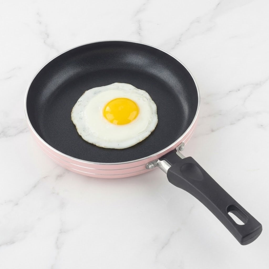 A pink frying pan with a black interior and handle holds a fried egg on a white marble-patterned surface.