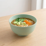 A light green speckled ceramic bowl filled with noodle soup, garnished with bok choy and carrot sticks, sitting on a light wood table.