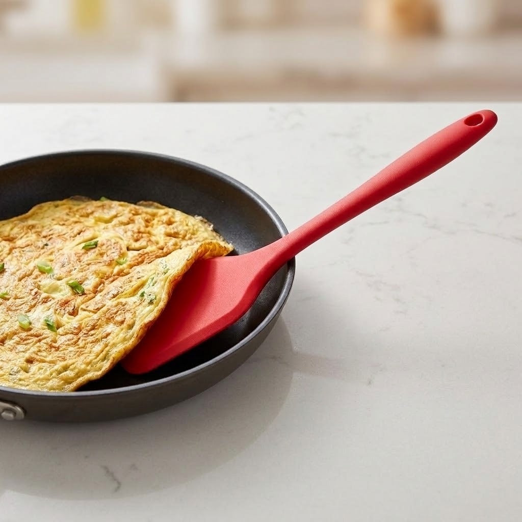 A red spatula slides beneath an omelette in a black frying pan on a pale countertop.