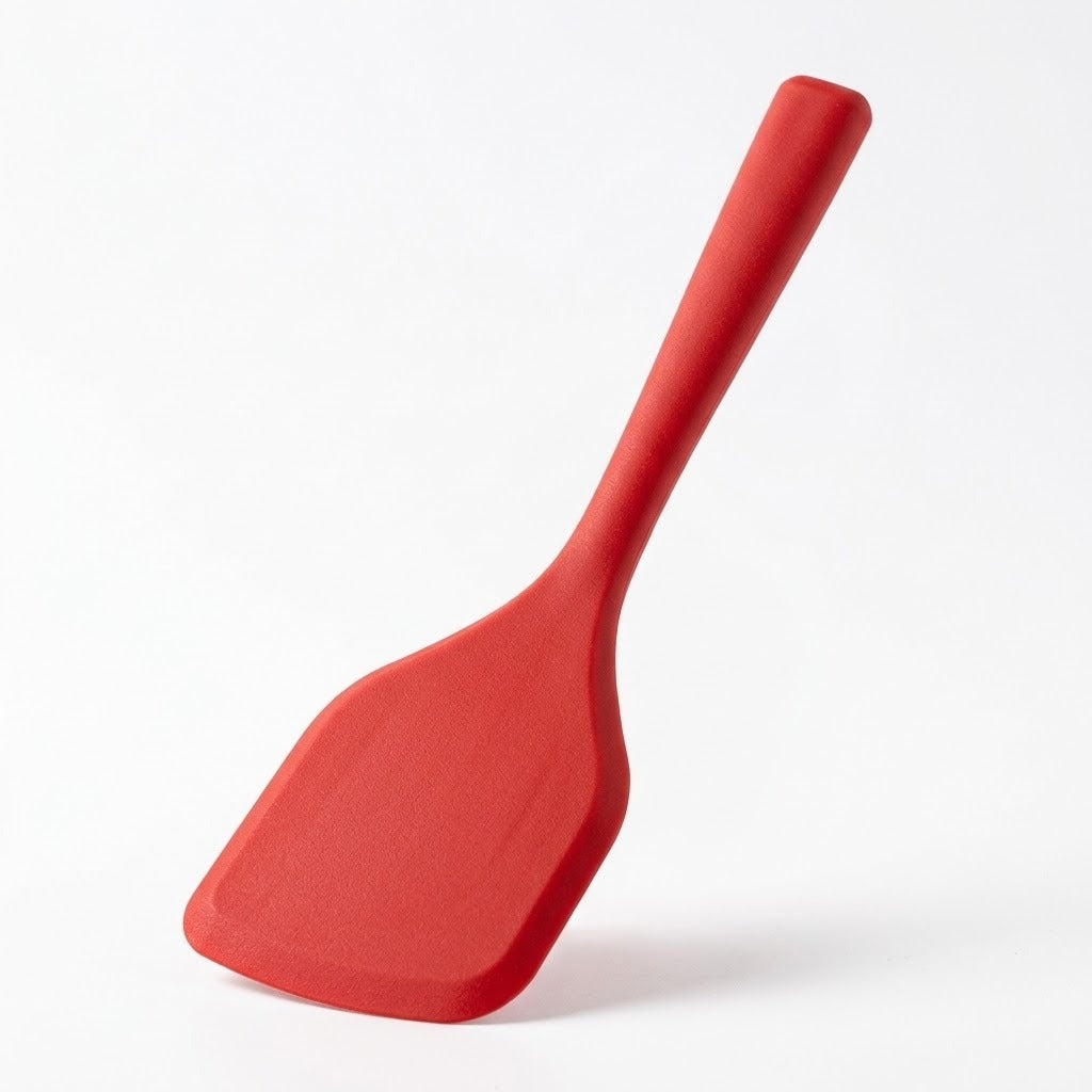 A red spatula stands angled on its broad head against a white background, casting a soft shadow.