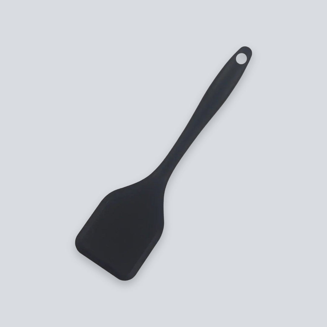 A black spatula with a broad head and round hanging hole lies diagonally against a light grey background.