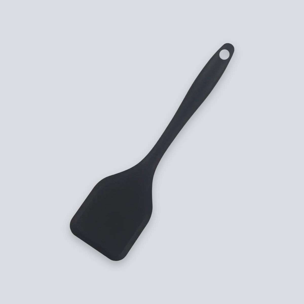 A black spatula with a broad head and round hanging hole lies diagonally against a light grey background.