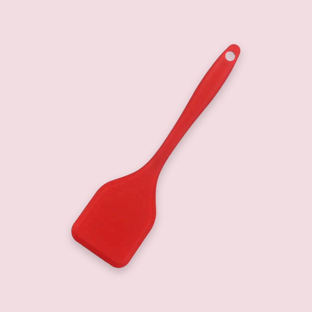 A red spatula with a broad head and round hanging hole lies diagonally against a pale pink background.