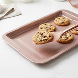 A rose-coloured spatula lifts a chocolate chip cookie from a matching baking tray on a white countertop.