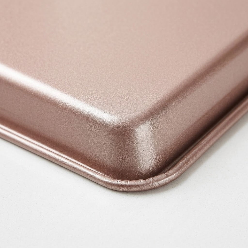 Close-up of a rose-coloured baking tray corner shows its finely textured surface and rolled rim against a white background.